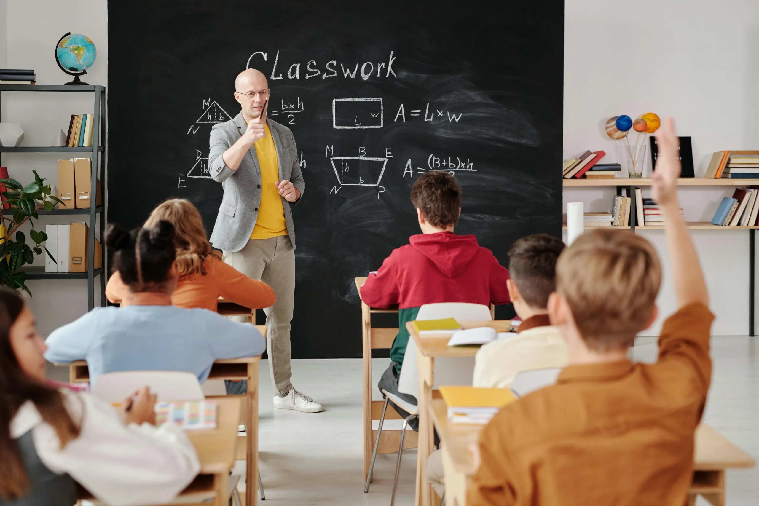 group of childrenin a c;assroom and a teacher 2 students are raising their hand.